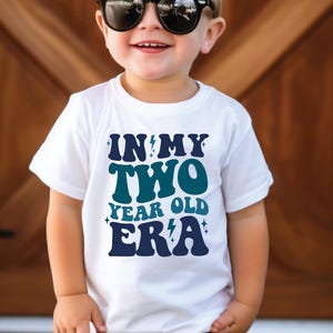 May include: White t-shirt with the text "IN MY TWO YEAR OLD ERA" in blue and teal. The shirt is worn by a young child wearing black sunglasses. The background is a wooden door.