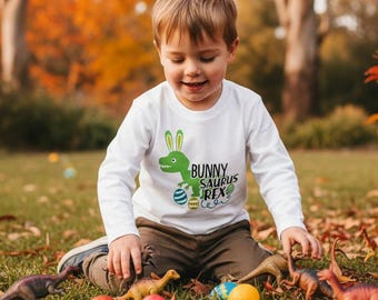 May include: A young child wearing a white long-sleeve shirt with a green dinosaur and bunny ears graphic that reads "Bunny Saurus Rex". The child is playing with toy dinosaurs and colorful Easter eggs on a grassy surface.