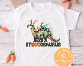May include: White t-shirt featuring a cartoon Stegosaurus wearing bunny ears and decorated with Easter eggs. The word "STEGGOSAURUS" is printed below the dinosaur. The shirt has a round neck and short sleeves. Carrots are placed around the shirt.
