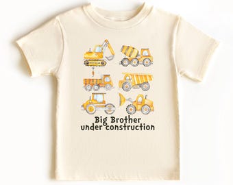 May include: A cream-colored short-sleeve t-shirt featuring a watercolor graphic of six yellow construction vehicles, including an excavator, cement mixer, and dump truck, with the black text "Big Brother under construction" printed below.