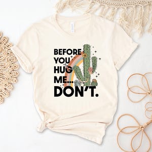 May include: Cream-coloured t-shirt with a cactus and rainbow graphic and the text "BEFORE YOU HUG ME... DON'T." The design includes a green cactus, a rainbow, and floral accents. The shirt is made of a soft material.