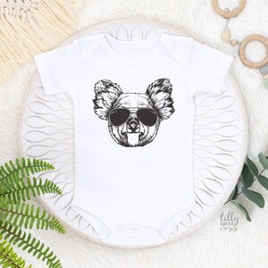 May include: A white short-sleeved baby bodysuit featuring a black and white line art graphic of a koala wearing aviator sunglasses centered on the chest. The garment has a round neckline and snap closures at the bottom for easy access.