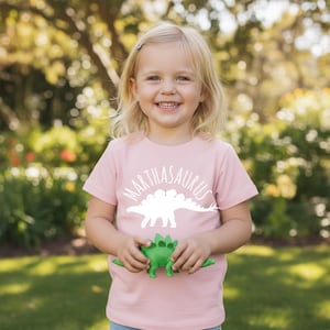 May include: A light pink short-sleeved t-shirt featuring a white graphic of a stegosaurus silhouette and the text "MARTHASAURUS" arched above it. The shirt is held by a small green plastic stegosaurus toy in the centre of the chest area.