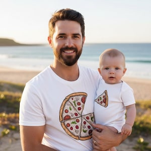 May include: A white short-sleeved t-shirt featuring a large graphic of a pizza with several slices, and a matching white short-sleeved baby bodysuit featuring a single graphic of a small pizza slice, both with pepperoni, mushroom, and basil toppings.
