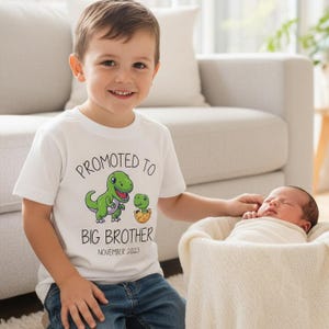 May include: A white short-sleeved t-shirt featuring a graphic of a large green dinosaur and a small green dinosaur hatching from an egg. The text reads PROMOTED TO BIG BROTHER and NOVEMBER 2023 in black lettering on the front of the garment.