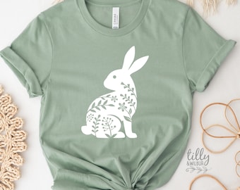 May include: A sage green t-shirt with a white floral bunny silhouette. The bunny is outlined with leaves and flowers. The text "tilly & wilbur" is in the bottom right corner.