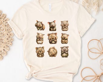 May include: A cream-coloured t-shirt displaying a grid of nine cartoon wombats in different poses. The wombats are rendered in shades of brown and tan, with expressive faces. The shirt has a classic crew neck and short sleeves.