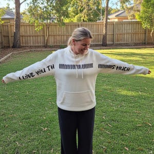 May include: Cream-coloured hoodie with black text on the sleeves and chest. The left sleeve reads "I LOVE YOU TH" and the right sleeve reads "IS MUCH". The chest features a series of vertical black lines. The hoodie has a front pocket and a hood.