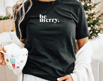 Könnte beinhalten: Ein schwarzes Kurzarm-T-Shirt mit dem weißen Schriftzug "be merry." auf der Brust. Das T-Shirt hat einen Rundhalsausschnitt und wird mit einer weißen Strickjacke sowie einer weißen Tasse mit Nussknacker-Motiv getragen.