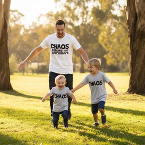 Tilly&Wilbur matching set — a white adult tee reading "Chaos Follows Me Everywhere" and grey t-shirts reading "Chaos" in bold rounded lettering, worn by a dad and two children playing outdoors