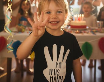 May include: A young child wearing a black t-shirt with a white hand graphic and the text "I'M THIS MANY". The child is smiling and holding up four fingers, celebrating a birthday. A birthday cake and party decorations are visible in the background.