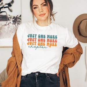 May include: A white short-sleeved t-shirt featuring the text "JUST ONE MORE" in stacked teal and orange retro-style lettering, with the word "chapter" written in a teal cursive script underneath. The shirt is worn with a brown button-down shirt.