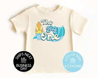 May include: A light beige t-shirt with a graphic of a surfboard with a sun and waves. The text "The Big One" is written in a retro font. The shirt has a black circle with the text "Ships Next Business Day" and a light blue circle with the text "Lots of Sizes & Colours".
