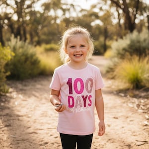 May include: A light pink short-sleeved t-shirt featuring the text 100 DAYS of school in glittery pink and purple gradient lettering. The number 100 is large and centred, with the word DAYS below it and the word of school in script.