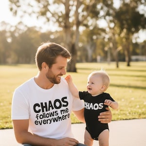 Tilly&Wilbur matching set — a white adult tee reading "Chaos Follows Me Everywhere" and a pblack baby bodysuit reading "Chaos" in bold rounded lettering, worn by a dad holding his laughing baby outdoors.