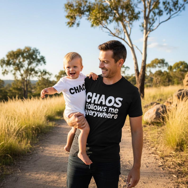 Tilly&Wilbur matching set — a black adult tee reading "Chaos Follows Me Everywhere" and a white baby bodysuit reading "Chaos" in bold rounded lettering, worn by a dad holding his laughing baby outdoors.