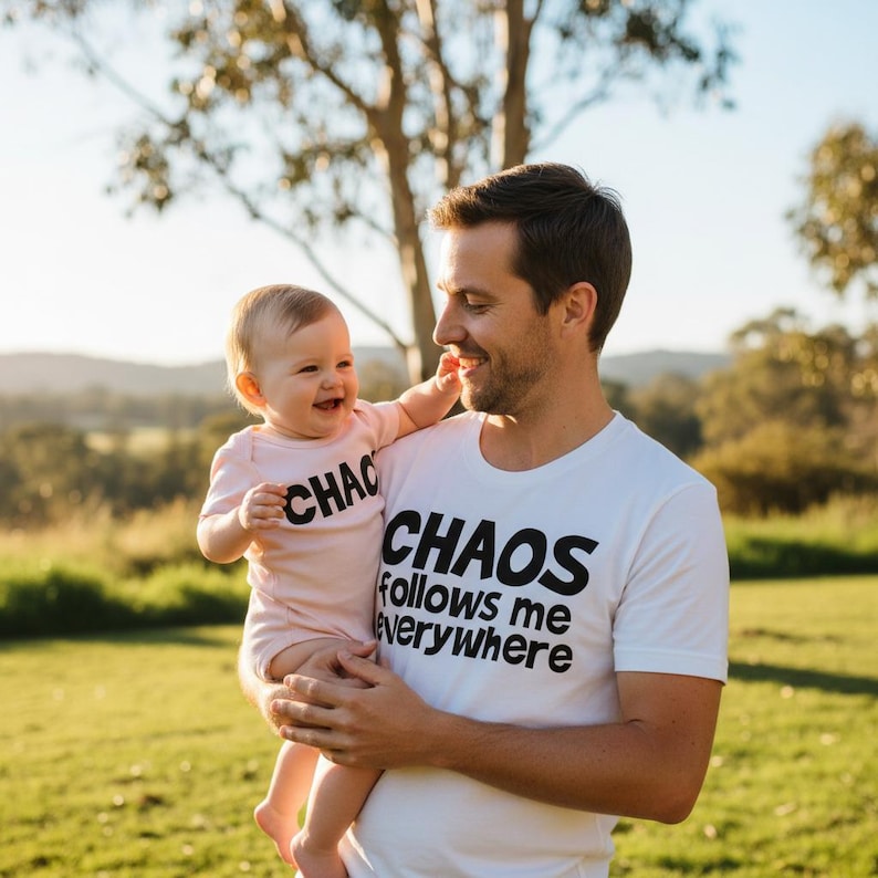 Tilly&Wilbur matching set — a white adult tee reading "Chaos Follows Me Everywhere" and a pink baby bodysuit reading "Chaos" in bold rounded lettering, worn by a dad holding his laughing baby outdoors.