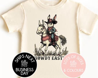May include: Cream-colored toddler t-shirt featuring a vintage-style illustration of a rabbit cowboy riding a horse. The text "HOWDY EAST" is printed on the shirt. Additional text on the shirt includes "SHIPS NEXT BUSINESS DAY" and "LOTS OF SIZES & COLOURS".