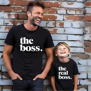 May include: Two black short-sleeved t-shirts with white text. One shirt features the text "the boss." and the other shirt features the text "the real boss." Both shirts have a crew neckline and are made of a solid black fabric with bold lettering.