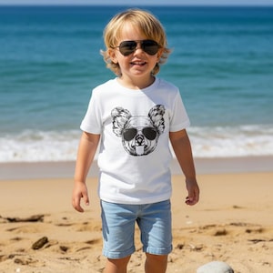 May include: A white short-sleeved t-shirt featuring a black and white graphic print of a koala wearing sunglasses on the chest. The shirt is paired with light blue denim shorts. The koala graphic mirrors the style of the sunglasses worn by the wearer.