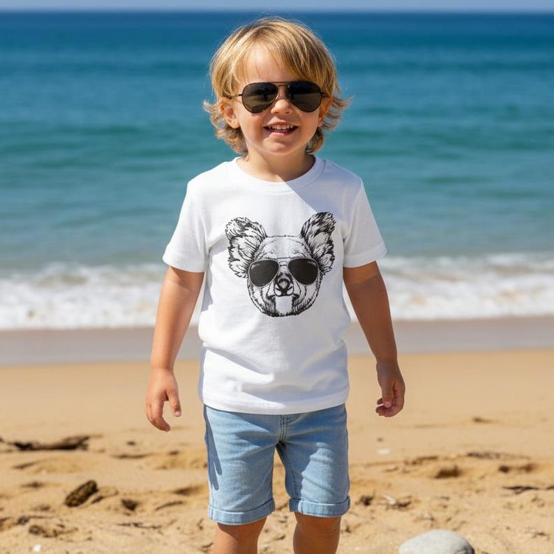 May include: A white short-sleeved t-shirt featuring a black and white graphic print of a koala wearing sunglasses on the chest. The shirt is paired with light blue denim shorts. The koala graphic mirrors the style of the sunglasses worn by the wearer.