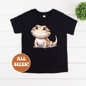 May include: Black t-shirt featuring a cartoon image of a smiling bearded dragon. The text "ALL SIZES!" is on a brown circle in the bottom left corner of the image.