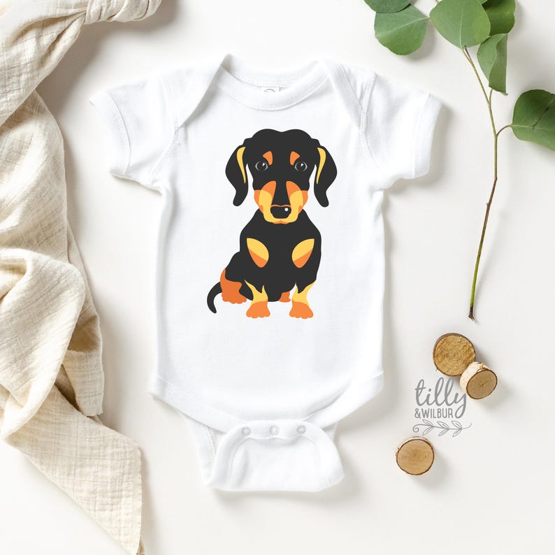 A white baby onesie with a black and orange dachshund dog graphic printed on it, along with some wooden coasters and a plant leaf.