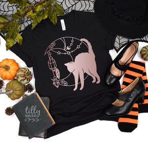 May include: Black t-shirt featuring a rose gold design of a cat silhouette on a crescent moon with stars, crystals, and a feather. The text "tilly & wilbur" is on a black book in the image.