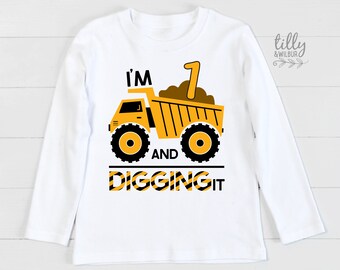 May include: White long-sleeved shirt featuring a yellow dumper truck graphic. The truck is carrying dirt and has the number "1" on it. The text "I'm 1 and digging it" is printed below the truck.