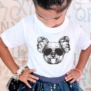 May include: A white short-sleeved t-shirt featuring a black and white line art graphic of a koala wearing aviator sunglasses. The text "tilly & wilbur" is printed in a cursive font on the left side of the shirt below the koala illustration.