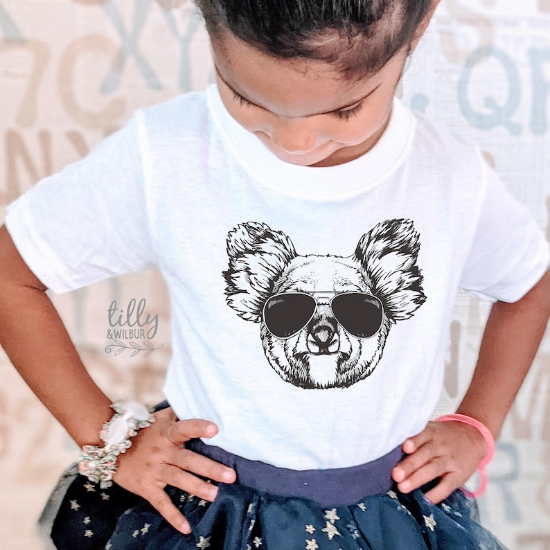 May include: A white short-sleeved t-shirt featuring a black and white line art graphic of a koala wearing aviator sunglasses. The text "tilly & wilbur" is printed in a cursive font on the left side of the shirt below the koala illustration.