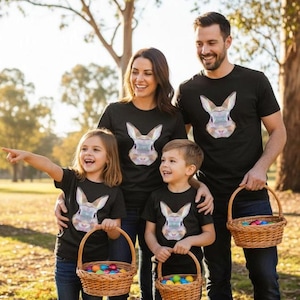 May include: A family of four wearing matching black t-shirts with a geometric rabbit design. Each person holds a wicker basket filled with colourful Easter eggs. The family is smiling and enjoying a sunny day outdoors.