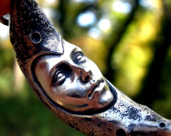 May include: A silver-toned metal pendant shaped like a crescent moon featuring a sculpted human face in the center. The surface of the moon has a textured, weathered finish with small circular indentations along the curved edges of the piece.