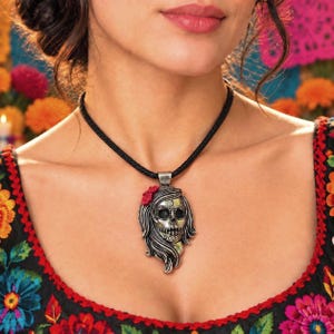 May include: A silver-toned pendant shaped like a stylized skull with flowing hair and a small red flower accent, suspended from a simple black braided cord necklace. The skull features intricate engraved details around the eyes and mouth area.