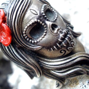 May include: A silver-toned Day of the Dead skull pendant with intricate detailing. The skull features decorative elements around the eyes and mouth, and a bright red flower adorns the side. The pendant has long, flowing hair.