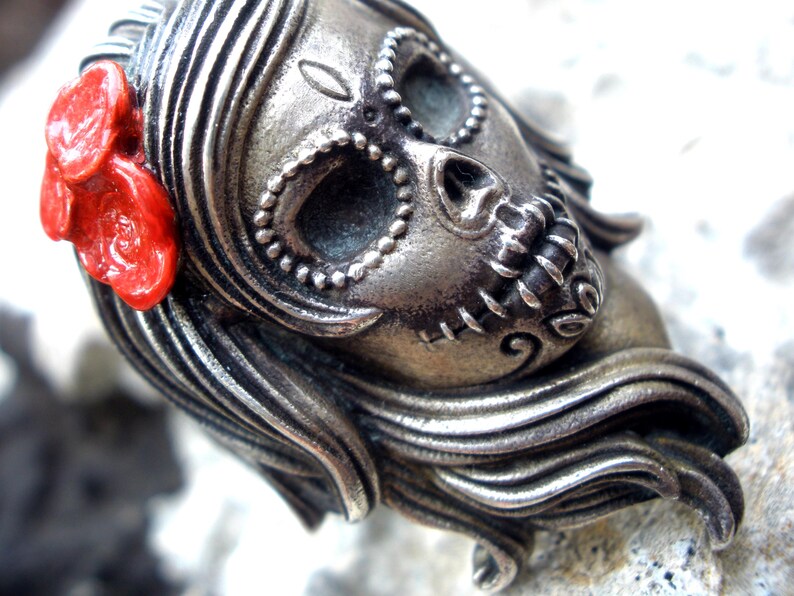 May include: A silver-toned Day of the Dead skull pendant with intricate detailing. The skull features decorative elements around the eyes and mouth, and a bright red flower adorns the side. The pendant has long, flowing hair.