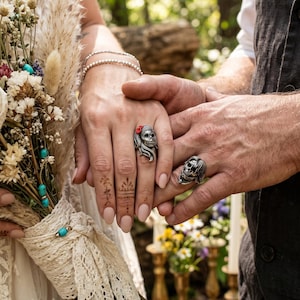 May include: Two silver skull rings, one with a red flower accent, are worn on the hands of a couple. The hands are clasped, with a bouquet of dried flowers and a lace-tied handle in the background. The setting appears to be outdoors.