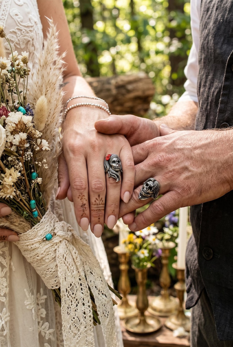 May include: Two silver skull rings, one with a red flower accent, are worn on the hands of a couple. The hands are clasped, with a bouquet of dried flowers and a lace-tied handle in the background. The setting appears to be outdoors.