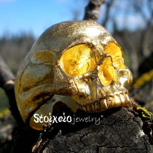 May include: A gold-colored ring shaped like a human skull with a textured surface and hollow eye sockets. The text Stoixeiojewelry is printed in white on the lower left side of the ring, which is positioned on a dark, weathered piece of wood.
