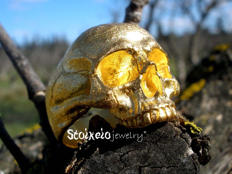 May include: A gold-colored ring shaped like a human skull with a textured surface and hollow eye sockets. The text Stoixeiojewelry is printed in white on the lower left side of the ring, which is positioned on a dark, weathered piece of wood.