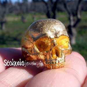 May include: A gold-colored ring featuring a detailed human skull design with a textured surface. The text Stoixeio jewelry is printed in white on the lower left side of the ring. The skull has hollow eye sockets and a defined nasal cavity.
