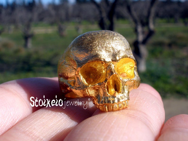 May include: A gold-colored ring featuring a detailed human skull design with a textured surface. The text Stoixeio jewelry is printed in white on the lower left side of the ring. The skull has hollow eye sockets and a defined nasal cavity.