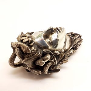 May include: A silver-toned metal pendant featuring a sculpted human face with closed eyes, surrounded by intricate, textured snake-like forms that wrap around the sides and back of the head, creating a detailed, three-dimensional metallic ornament.