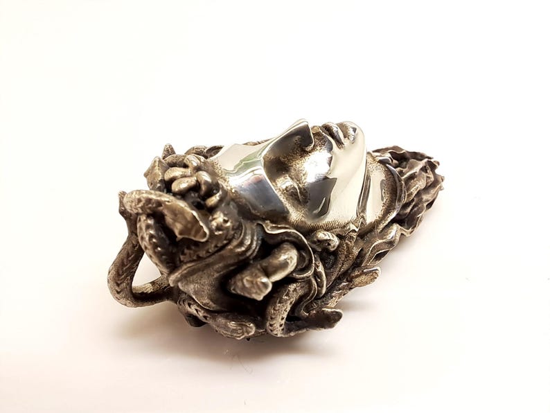 May include: A silver-toned metal pendant featuring a sculpted human face with closed eyes, surrounded by intricate, textured snake-like forms that wrap around the sides and back of the head, creating a detailed, three-dimensional metallic ornament.