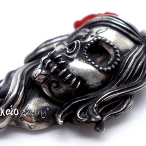 May include: A detailed silver-toned pendant in the shape of a stylized skull with long hair and a red flower. The skull has intricate detailing, including a beaded necklace and decorative elements. The pendant is likely jewelry.