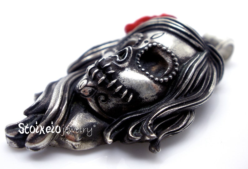 May include: A detailed silver-toned pendant in the shape of a stylized skull with long hair and a red flower. The skull has intricate detailing, including a beaded necklace and decorative elements. The pendant is likely jewelry.