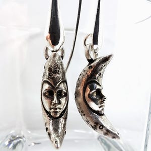 May include: A pair of silver-toned drop earrings featuring a crescent moon design. Each earring displays a detailed human face sculpted within the curve of the moon. The metal has a textured, weathered finish with polished highlights on the facial features.