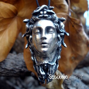 May include: A silver-toned pendant featuring a sculpted human face with closed eyes and snakes intertwined in the hair, suspended from a black cord. The text Stoixeio jewelry is printed in white in the lower right corner of the image.