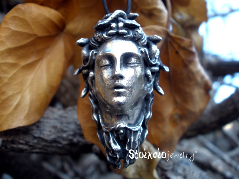 May include: A silver-toned pendant featuring a sculpted human face with closed eyes and snakes intertwined in the hair, suspended from a black cord. The text Stoixeio jewelry is printed in white in the lower right corner of the image.