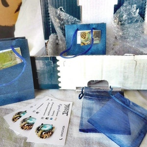 May include: An assortment of packaged jewelry items. The image shows blue gift bags, small blue drawstring pouches, and business cards with a logo and contact information. The items are arranged in a cardboard box with bubble wrap.